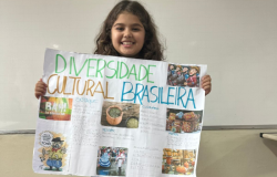 #paratodosverem: na imagem, uma menina sorridente segura um cartaz colorido com o título "Diversidade Cultural Brasileira", que mostra imagens e textos sobre sotaques, culinária, festas e artes do Brasil. Ao fundo, há um quadro branco de sala de aula.