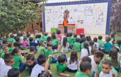 #paratodosverem: Crianças de camiseta verde estão sentadas na grama, em uma grande roda, assistindo a uma contadora de histórias ou professora. A mulher, de blusa preta e avental laranja, está em pé em frente a um mural cheio de desenhos e recados.