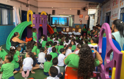 #paratodosverem: Em uma sala de aula colorida, com um grande brinquedo infantil, várias crianças de camiseta verde estão sentadas no chão em semicírculo, voltadas para uma tela digital. A tela exibe imagens de árvores. Duas professoras estão com o grupo. A cena, vibrante e cheia de cores, mostra um momento de aula interativa e lúdica.