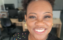 Mulher em sala com computadores ao fundo, sorri para foto