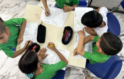 A imagem mostra um grupo de cinco crianças reunidas em torno de uma mesa em uma sala de aula. Elas estão vestindo camisetas verdes, com exceção de uma criança que veste branco, como parte de um uniforme escolar. As crianças estão realizando uma atividade educacional, escrevendo e colorindo com lápis de cor e lápis grafite. Elas parecem concentradas e envolvidas na atividade, compartilhando materiais como estojos e lápis. O ambiente possui cadeiras azuis, uma mesa bege e um piso de mármore branco com veios e