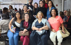 #paratodosverem: Um grupo de pessoas posa sorridente para a foto em um saguão iluminado. Algumas estão sentadas em um banco e outras atrás, em pé atrás, segurando bolsas e ingressos. Ao fundo há mais pessoas reunidas e uma placa indicando “Grande Teatro Cemig Palácio das Artes”