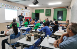 #paratodosverem: a foto mostra um educador na frente de um quadro, em uma sala pintada de bege, com janelas e portas verdes. Acima do quadro, há papéis com as letras do alfabeto. No teto, um projetor exibe uma apresentação. As carteiras estão quase todas ocupadas com pessoas adultas de diversas idades. Algumas usam uniformes e estão prestando atenção no que o professor explica.
