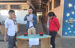 #paratodosverem: Na foto, dois estudantes participam de um jogo de perguntas e respostas em uma escola, enquanto um professor conduz a atividade com um microfone e uma folha de anotações. À frente deles há uma mesa com botões vermelhos, simulando um quiz. O clima é de concentração e aprendizado em um ambiente descontraído e educativo.