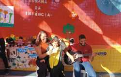 #paratodosverem: em frente a um painel laranja decorado com flores e brinquedos, uma criança fantasiada de dinossauro canta sorridente ao microfone, acompanhada por uma mulher ao lado e um homem tocando violão. A cena faz parte da “Semana da Infância”.