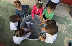 #paratodosverem: a foto mostra crianças pequenas ao redor de uma grande bacia cheia de água, brincando com barquinhos feitos de papel. Elas observam, atentas, os barquinhos boiando, enquanto algumas colocam as mãos na água. 
