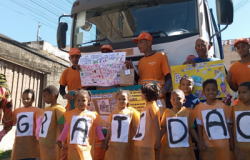 #paratodosverem: Um grupo de crianças veste aventais laranjas com letras que formam a palavra “GRATIDÃO”, posando em frente a um caminhão branco. Atrás delas, garis uniformizados de laranja seguram cartazes coloridos com mensagens de agradecimento e desenhos. A cena acontece em uma rua, com céu azul e fiação elétrica visível ao fundo.