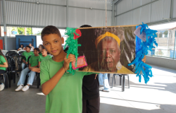 #paratodosverem: a imagem mostra, em um espaço coberto e fechado nas laterais por grades, um menino com uniforme escolar verde segurando; à sua direita, uma foto impressa de Conceição Evaristo colada em um cartaz com papel crepom verde e azul. Ao fundo, outros(as) estudantes uniformizados(as) estão sentados(as) em cadeiras de plástico brancas.