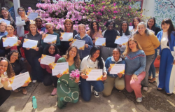 #paratodosverem: Um grupo de mulheres posa sorridente, ao ar livre, segurando certificados com bordas floridas. Atrás delas, há arbustos floridos de cor rosa e uma parede colorida com desenhos de animais. Algumas estão agachadas na frente, enquanto outras permanecem em pé, formando uma grande fileira. 