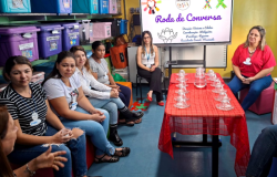 #paratodosverem: a foto mostra uma roda de conversa realizada em uma sala com parede amarela e uma estante com caixas organizadoras, à esquerda. Em frente à estante, algumas mulheres estão sentadas. No centro da sala, há uma mesa coberta com toalha vermelha, com várias xícaras de vidro e uma jarra com água. Ao fundo, um monitor exibe a arte gráfica do evento com o título “Roda de Conversa”.