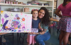 #paratodosverem: na imagem, em primeiro plano, mãe e filha estão em uma sala com paredes brancas e chão verde musgo. A mãe está agachada, à altura da criança, vestida com calça jeans azul e blusa verde. A criança usa um vestido azul marinho, branco e rosa. A mãe segura um quadro multicolorido pintado pela filha, com a presença de flores e mãos. Ao fundo, há prateleiras brancas com materiais pedagógicos, como papéis e recipientes cheios de lápis de cor. À direita, estão duas mulheres adultas.