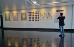 #paratodosverem: foto de uma sala de galeria com piso preto reflexivo. Em uma parede branca, são exibidas obras de arte com influência indígena: quadros geométricos coloridos, balaios com padrões tribais e peças artesanais penduradas. Um homem de costas fotografa o cartaz explicativo na parede iluminada.