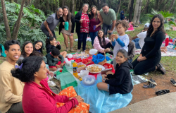 #paratodosverem: a imagem mostra, em um parque, um grupo de pessoas, incluindo pessoas adultas e crianças, reunido num piquenique em meio à natureza. Todos(as) estão em clima de alegria e descontração, alguns(mas) sentados(as) ao redor de uma toalha, sobre a qual há diversas guloseimas e bebidas, enquanto outros(as) estão em pé, ao fundo, sorrindo para a foto. O ambiente é arborizado, com bastante vegetação.