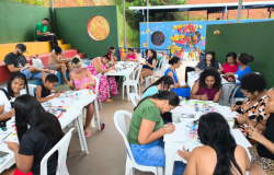 #paratodosverem: mulheres, crianças e alguns homens participam de uma atividade coletiva de pintura em telas, com mesas cheias de tintas, pincéis e telas, em um ambiente colorido, com a parte superior das paredes aberta ao exterior. Ao fundo da imagem, há brinquedos em uma área externa e, atrás deles, um muro azul com desenhos. Mais além, está uma área com plantas. 