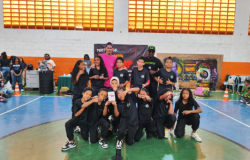 #paratodosverem: Um grupo de crianças e adolescentes veste roupas pretas iguais e posa no centro de uma quadra esportiva, alguns agachados e outros em pé, fazendo gestos com as mãos. Atrás deles, dois adultos estão de pé, um de rosa e outro de boné verde com microfone. No fundo há um banner “Festival Hip Hop”, grafite colorido e pessoas assistindo. Alguns estudantes na frente seguram um prêmio em formato de um retângulo branco.#paratodosverem: Um grupo de crianças e adolescentes veste roupas pretas iguais e