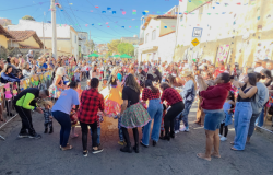#paratodosverem: na foto, pessoas adultas caracterizadas para uma festa junina, em roda, seguram a barra do vestido de uma garota que está no centro, em uma rua enfeitada com bandeirinhas coloridas. Ao redor, familiares observam e registram o momento com celular. O céu está azul e o dia ensolarado. Grades decoradas com temas juninos cercam o espaço.