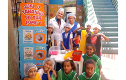 #paratodosverem: na foto, crianças posa ao lado de uma profissional de jaleco branco. À esquerda, um painel colorido exibe a pergunta: “Qual o tamanho da sua fome?” As crianças estão organizadas junto a uma mesa com caixas plásticas e bacias de metal. Algumas usam coletes coloridos e toucas. Ao fundo, vê-se uma escada verde e parte da área externa da escola.