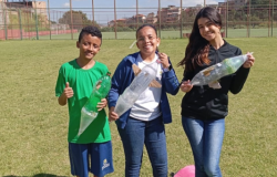 #paratodosverem: Três estudantes estão em um campo de futebol gramado, sorrindo e segurando foguetes feitos com garrafas PET. O aluno à esquerda veste camiseta verde e bermuda azul. Ao centro, uma aluna de óculos usa jaqueta azul. À direita, outra aluna com cabelos longos veste jaqueta preta. O céu está azul e há casas ao fundo.