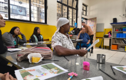 #paratodosverem: em uma sala de aula com paredes amarelas e janelas gradeadas, um grupo de pessoas adultas está sentado ao redor de mesas, prestando atenção a alguém fora de cena. Sobre a mesa, há panfletos coloridos com o título “Arte e terapia”, lápis, apontador, uma caneca metálica e um pedaço de bolo em um guardanapo. Ao fundo, um homem de blusa marrom está em pé.