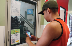 #paratodosverem: em um corredor, um homem com boné e camiseta vermelha e preta lê o QR Code em um panfleto colado em uma porta; ele está virado de lado para a câmera, com seu celular apontado para o panfleto.