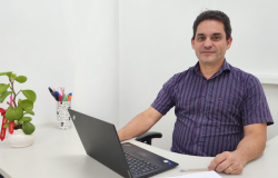 Homem branco, sentado à mesa, usando camisa social listrada com tons em azul e roxo, utilizando notebook. 