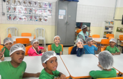 #paratodosverem: Em uma sala de aula clara, com mesas e cadeiras laranja e brancas, um grupo de crianças está sentado em torno de duas mesas. Todas usam toucas brancas de cabelo e camisetas de uniforme. Algumas sorriem para a câmera, exibindo entusiasmo em um momento de preparo para uma atividade.