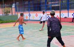 #paratodosverem: Fotografia colorida de três crianças jogando futebol na quadra da escola. Duas crianças estão de costas para a câmera e uma está de frente, chutando a bola.