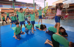 #paratodosverem: na foto, crianças uniformizadas realizam uma apresentação de ginástica em grupo sobre um tatame azul em uma quadra coberta. Elas formam pirâmides humanas. Outras crianças assistem ao fundo, com duas mulheres. A maioria dos(as) estudantes veste camiseta verde e bermuda azul.