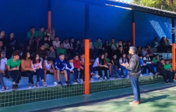 #paratodosverem: A imagem mostra um grupo grande de estudantes sentados em arquibancada coberta, dividida em dois níveis, com paredes azuis e colunas laranja. Eles observam atentamente um homem de jaqueta preta e calça jeans que está em pé na frente, gesticulando enquanto fala. O espaço é iluminado pela luz natural e tem piso verde.