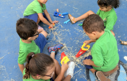 #paratodosverem: Cinco crianças sentadas no chão azul participam de uma atividade. Elas usam uniformes verdes e exploram ferramentas de brinquedo enquanto manuseiam palhas espalhadas pelo chão. Uma menina de cabelo preso em rabo de cavalo baixo e cacheado observa, enquanto outras, com cabelos soltos ou presos, interagem em grupo, formando uma roda.