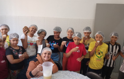 #paratodosverem: Na imagem, um grupo de crianças posa sorridente em uma cozinha, todas usando toucas de proteção. Elas seguram copinhos com suco e demonstram alegria pela atividade. Uma mulher, também de touca, está sentada à frente, segurando uma jarra com a bebida, enquanto uma das crianças exibe um abacaxi. A cena transmite aprendizado e diversão em um momento coletivo.