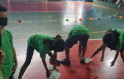 #paratodosverem: Na foto, um grupo de crianças participa de uma brincadeira no ginásio da escola. Usando vendas nos olhos e camisetas verdes, elas se abaixam tentando encontrar objetos no chão, guiando-se apenas pelo tato e pelas orientações dos colegas. A atividade promove cooperação, confiança e diversão em um ambiente educativo e acolhedor.