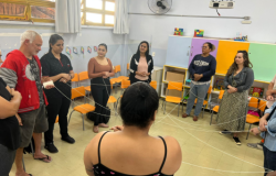 #paratodosverem: Em uma sala de aula, um grupo de adultos está em pé, formando um círculo. Eles seguram fios de barbante, criando uma teia entre si, como parte de uma atividade coletiva. Ao fundo, há cadeiras laranjas encostadas na parede, armários coloridos e materiais pedagógicos. A sala é iluminada por janelas e há um ventilador na parede.