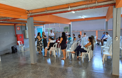 A imagem mostra um espaço com várias cadeiras brancas onde educadores(as) estão reunidos(as). Alguns(algumas) estão sentados(as) e outros(as) em pé. 