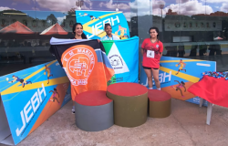 #paratodosverem: a imagem mostra uma cerimônia de premiação dos Jogos Escolares, realizada em um ambiente ao ar livre, em frente a um prédio com fachada de vidro espelhado. Três jovens atletas estão posicionadas sobre um pódio. As atletas que conquistaram o primeiro e o segundo lugar seguram as bandeiras de suas escolas: Professor Daniel Alvarenga e Marlene Pereira Rancante, respectivamente.