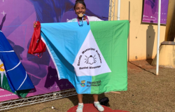 #paratodosverem: Uma menina está em um pódio vermelho segurando uma bandeira com as cores azul, branca e verde. Na bandeira está escrito “Escola Municipal Professor Daniel Alvarenga” e “Prefeitura Belo Horizonte”, além de um sino desenhado. Ela morde uma medalha dourada pendurada no pescoço e segura uma bolsa vermelha.