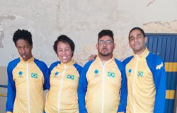 #paratodosverem: quatro pessoas estão lado a lado, vestindo agasalhos esportivos nas cores azul e amarelo, com a bandeira do Brasil no peito. Eles posam sorridentes para a foto, transmitindo união e orgulho. Ao fundo, uma parede com pintura desgastada e uma porta azul.