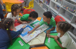 #paratodosverem: na foto, crianças uniformizadas estão reunidas ao redor de uma mesa colorida, interagindo com um livro coletivo cheio de desenhos e frases. As mesas e cadeiras são em tons de verde, vermelho, amarelo e azul. Ao fundo, prateleiras brancas organizadas guardam brinquedos e materiais, que estão dentro de caixas transparentes.