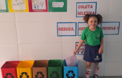 paratodosverem: na foto, uma criança uniformizada sorri ao lado de lixeiras coloridas de coleta seletiva, identificadas como “plástico”, “metal”, “orgânico”, “vidro” e “papel”. Ela deposita uma folha na lixeira azul, destinada ao papel. Ao fundo, cartazes educativos reforçam a importância da reciclagem com os dizeres "Reduza, Reuse, Recicle!"