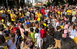 #paratodosverem: crianças e pessoas adultas estão em um evento ao ar livre. As crianças estão reunidas no centro, muitas segurando flores amarelas, enquanto as outras pessoas formam um círculo ao redor, demonstrando apoio.