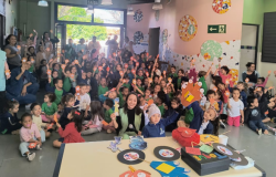 #paratodosverem: na foto, um grande grupo de crianças está reunido em um salão escolar, acompanhado por educadores(as). Muitas delas seguram palitinhos com mãos coloridas de papel. No ambiente, há decorações coloridas nas paredes, com mosaicos e placas de sinalização. À frente, uma mesa exibe discos de papel e materiais de artesanato.