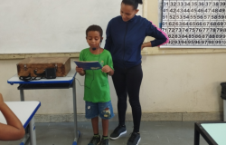 #paratodosverem: Um menino de uniforme verde e bermuda jeans lê um livro em voz alta, apoiado por um microfone de cabeça. Ele está no centro de uma sala de aula e, ao lado dele, uma professora o observa com carinho. A cena mostra um momento de leitura e apresentação oral em frente a um quadro branco e um cartaz numérico.