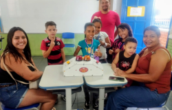 Em uma sala de aula, duas pessoas adultas sentam-se uma de frente para a outra, com duas mesas entre elas. Em cima destas, há uma máquina com um botão e uma sirene de cada lado, usada para jogos de pergunta e resposta. Em volta das mesas, há 5 crianças e um adulto sorrindo para a foto. Na parede ao fundo, está afixado um quadro branco e, ao lado dele, há uma porta de cor azul. A parede abaixo do quadro está pintada na cor verde. 