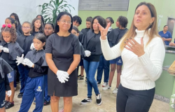 #paratodosverem: na imagem, ao centro, uma mulher de vestido preto também usa luvas, e ao lado dela uma mulher de blusa branca de gola alta gesticula enquanto fala. Ao fundo, um grupo de crianças e adolescentes está reunido. Eles vestem uniforme azul com sobreposição preta e usam luvas brancas