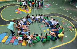 #paratodosverem: A imagem mostra um grupo de crianças e adolescentes sentados(as) no chão de um pátio escolar. Eles(as) estão vestindo uniformes escolares, predominantemente nas cores azul e verde, e alguns(mas) estão usando meias coloridas e descombinadas, criando um visual divertido e chamativo. Algumas pessoas adultas também estão presentes, ajudando na atividade. O chão do pátio tem desenhos coloridos de jogos tradicionais, como amarelinha e twister.