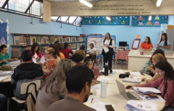 #paratodosverem: na foto, em uma biblioteca escolar, vários(as) educadores(as) estão sentados(as) em mesas redondas, concentrados(as) em leituras e anotações. Ao fundo, uma mulher conduz a apresentação em pé, segurando papéis. Atrás dela, há uma mesa com garrafa de café e um lanche. Outras pessoas estão próximas ao balcão da biblioteca. À esquerda, há estantes repletas de livros. 