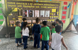 #paratodosverem: na imagem, crianças uniformizadas observam com atenção um mural escolar com o cartaz: "Vamos celebrar a resistência, resiliência e a beleza das tradições dos povos indígenas!". O painel conta com cartazes informativos, objetos artesanais e elementos decorativos, como penas e palha, criando um ambiente de aprendizado visual e interativo.