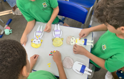#paratodosverem: na imagem, quatro crianças, com camisetas verdes do uniforme escolar, estão reunidas em torno de uma mesa branca. Sobre ela, há bonecos(as) de papel e círculos adesivos coloridos, representando o “Semáforo dos Toques". As crianças colam os adesivos nos(as) bonecos(as). Ao fundo, há cadeiras azuis e parte do ambiente escolar.