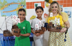 #paratodosverem: a imagem mostra três pessoas em destaque: duas alunas e uma professora, todas segurando maquetes de caravelas portuguesas feitas com material reciclável. As alunas usam uniformes (uma verde, outra branco) e a professora veste blusa amarela, avental com estampa de abacaxi e enfeite de abacaxi na cabeça. Ao fundo, há uma parede de azulejos brancos e alaranjados, com um mapa e uma maquete do mar com uma caravela.