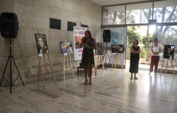 #paratodosverem: A imagem mostra a entrada da Smed, onde uma mulher segura um microfone e um panfleto enquanto discursa, cercada por obras fotográficas expostas em cavaletes. Duas outras mulheres acompanham a apresentação, uma de braços cruzados e outra segurando papéis. O ambiente conta com grandes portas de vidro que permitem a entrada de luz natural. Ao fundo, aparecem outras plantas em vasos e algumas árvores.